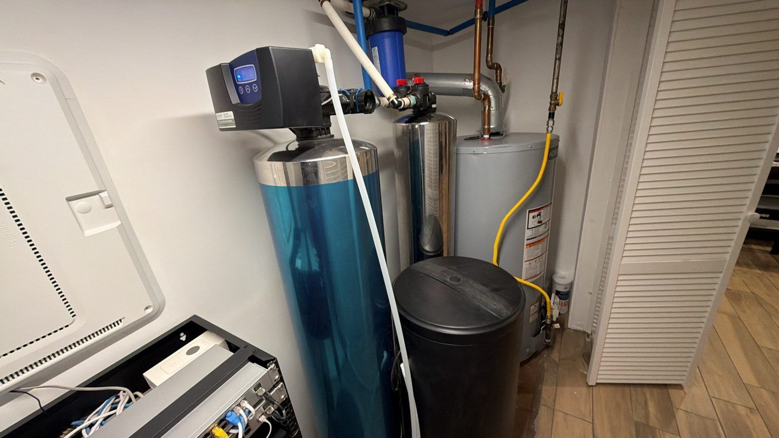 The digital control valve on top of a blue water softener tank, with a stainless carbon filter tank, a gas water heater and the black salt tank behind and beside it in a utility closet.