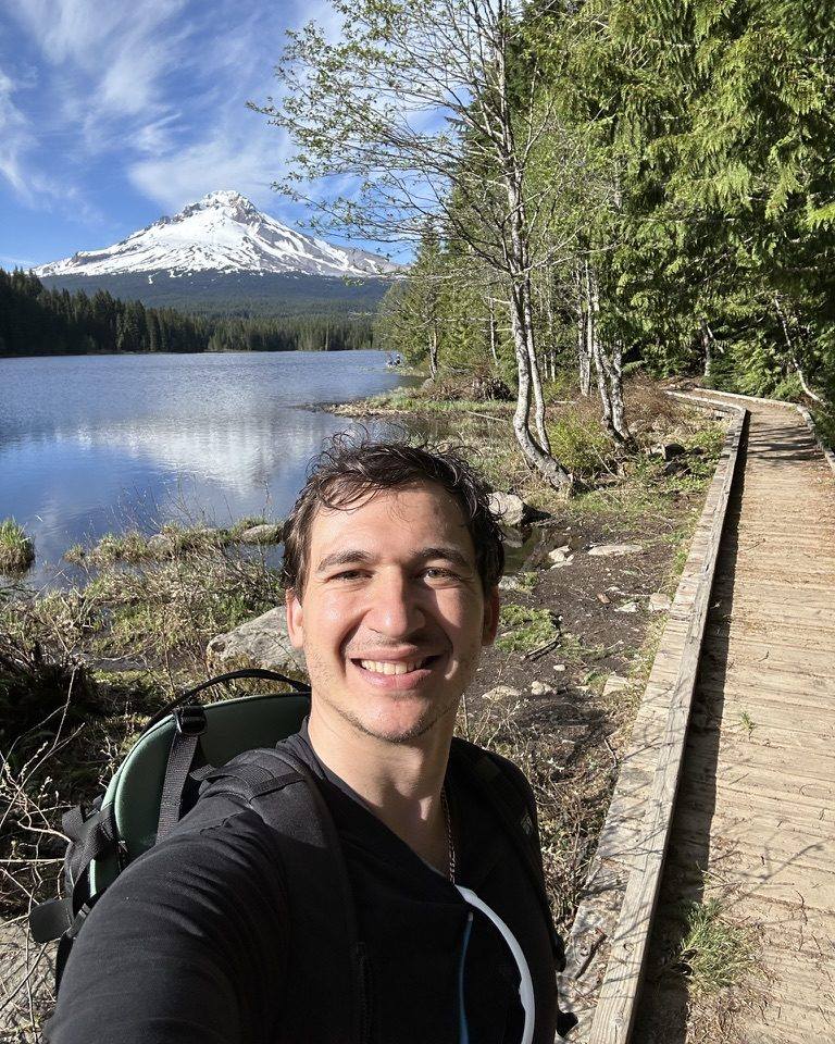 Dom, the owner, smiling outdoors on a lakeside trail